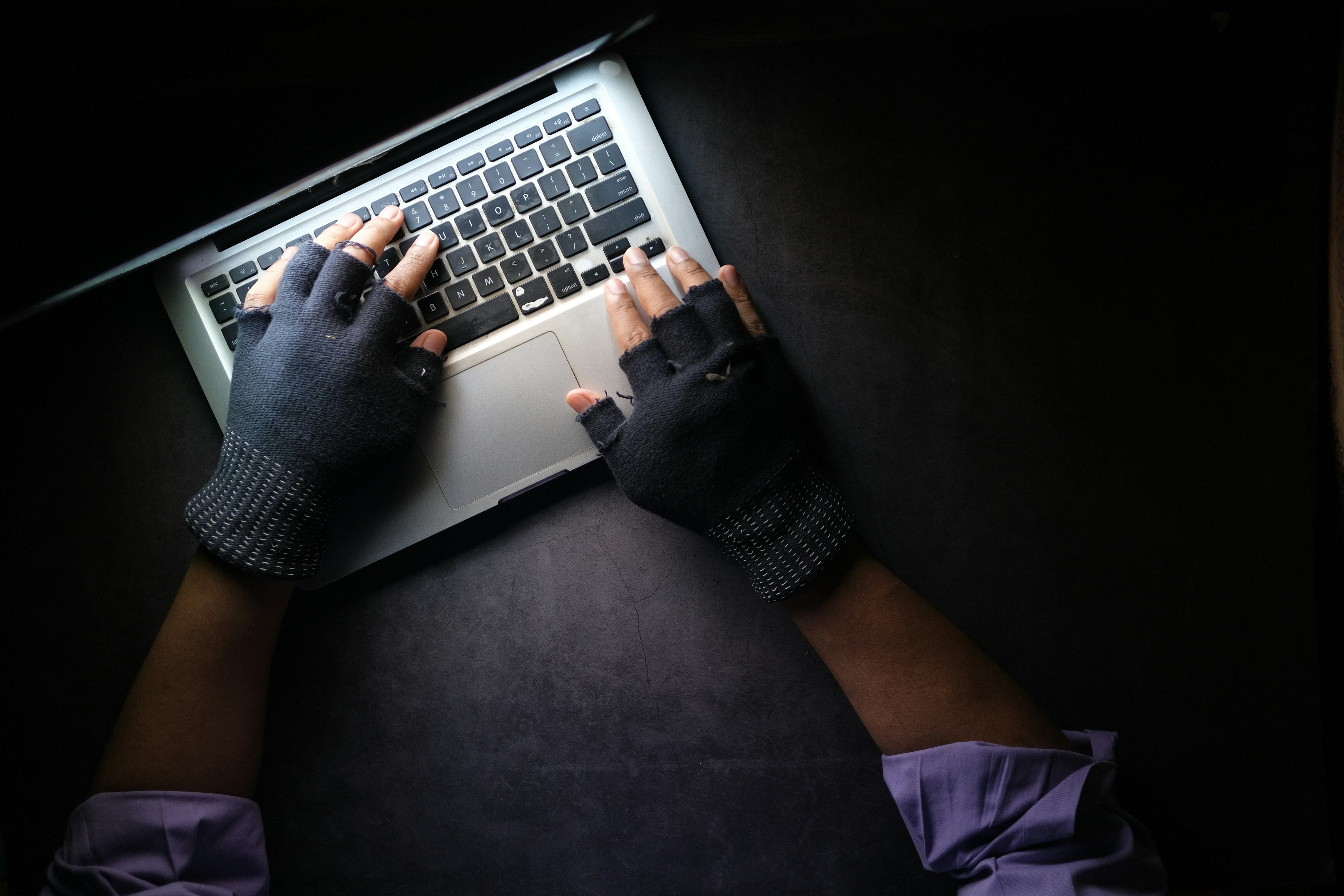A person wearing fingerless gloves typing on a laptop on a dark surface